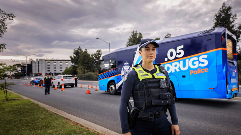 A police officer stands in front of a blue roadside drug testing bus, reflecting Victoria's Driving Law Reform. Orange cones line the road, highlighting active enforcement measures in the background.