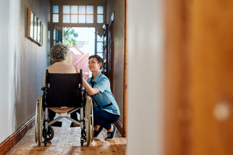 A caregiver kneels while speaking to an elderly person seated in a wheelchair in a well-lit hallway, demonstrating their understanding and dedication to new quality standards in aged care.