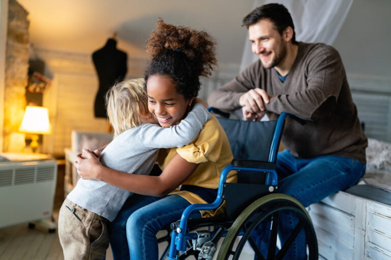 A young girl in a wheelchair is being hugged by a young boy while a man sits nearby and smiles, reflecting the changes supported by NDIS.