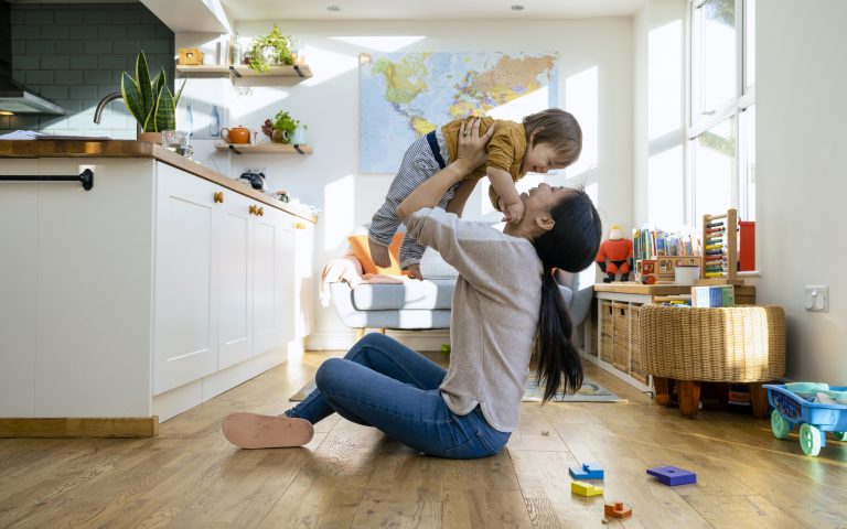 A woman sits on a wooden floor, lifting a toddler in the air in a bright, cozy living room with a kitchen and toys around, embodying the joy found even amidst the ongoing child care crisis.