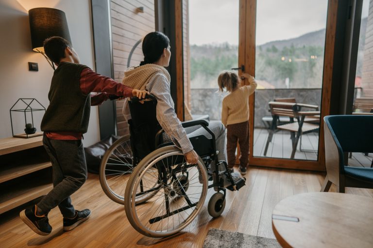 A person in a wheelchair is pushed by a child, while another child looks out a glass door onto the balcony. The room, with its wooden floors and view of trees and hills, seems to reflect one of the key updates highlighted in the 2023-24 Annual Report.