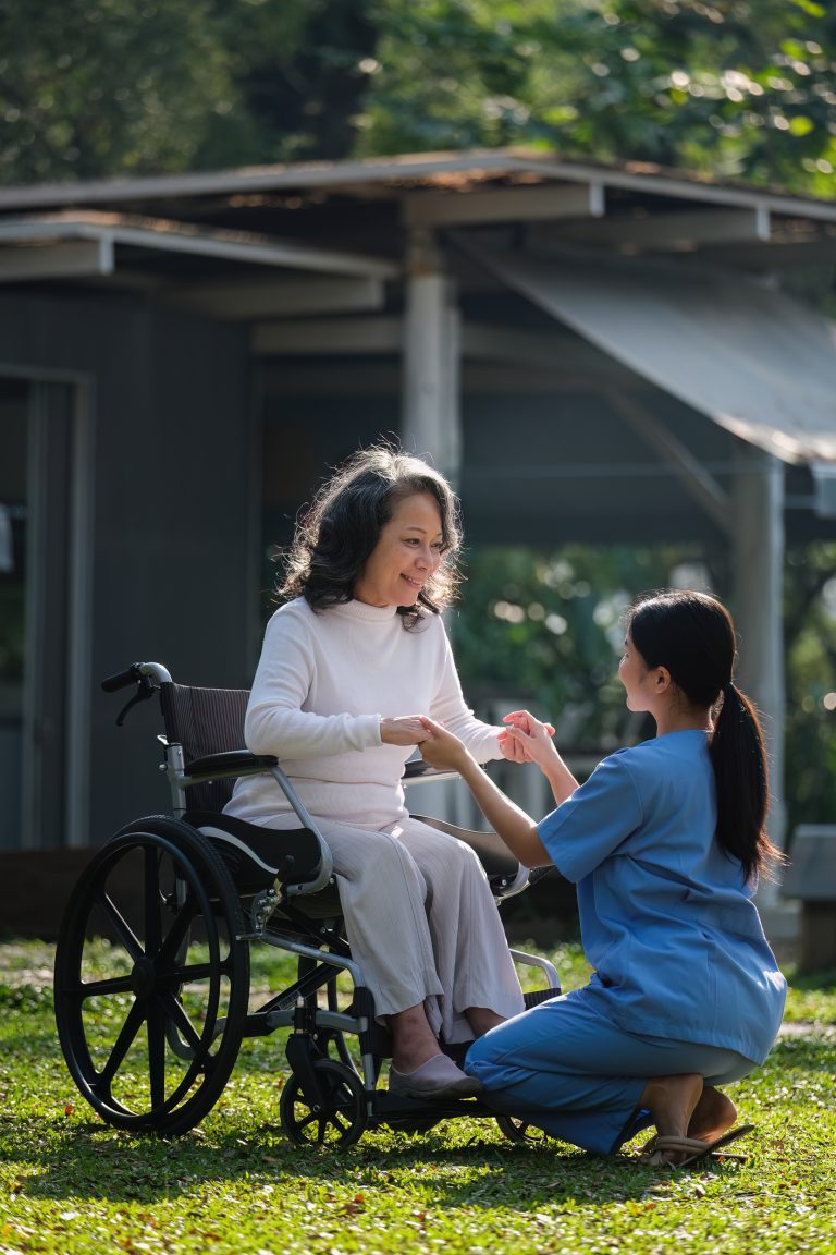 A woman in a wheelchair holds hands with a nurse in blue scrubs outdoors on a sunny day, highlighting the government's commitment to improving home care for older Australians, set against the backdrop of a small building.