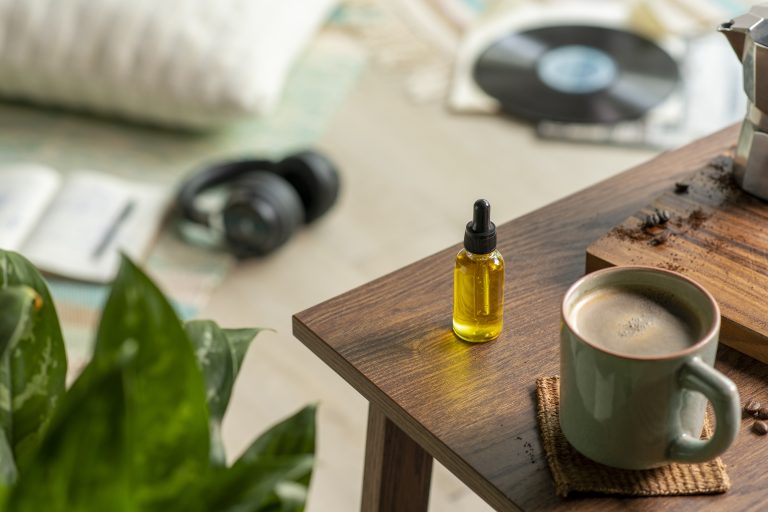 A dropper bottle of oil and a mug sit on a wooden table. In the blurred background, headphones, a vinyl record, and a plant hint at serenity. The scene captures the soothing boom of medicinal cannabis culture emerging in Australia.