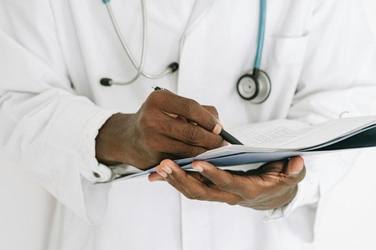 A person in a white lab coat with a stethoscope writes on a clipboard.
